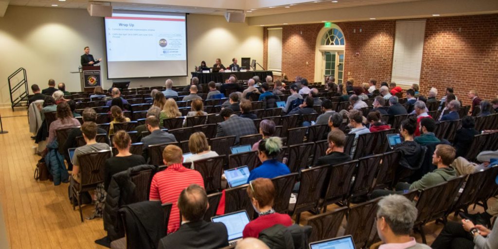 Auditorium space with rows of people and a presenter at the front of the room standing next to a screen