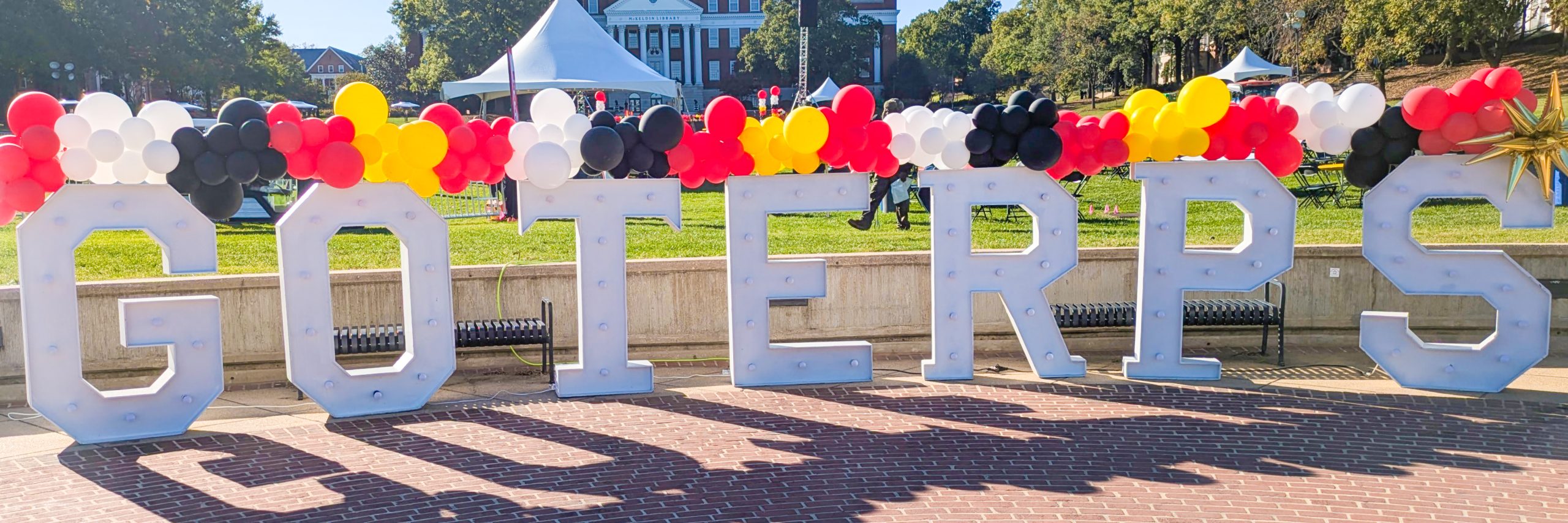 Large, white, marquee-style letters spelling out "GO TERPS" are decorated with an arc of red, black, yellow, and white balloons. The letters are set up on a brick plaza on a sunny day, with campus buildings and white tents visible in the background.