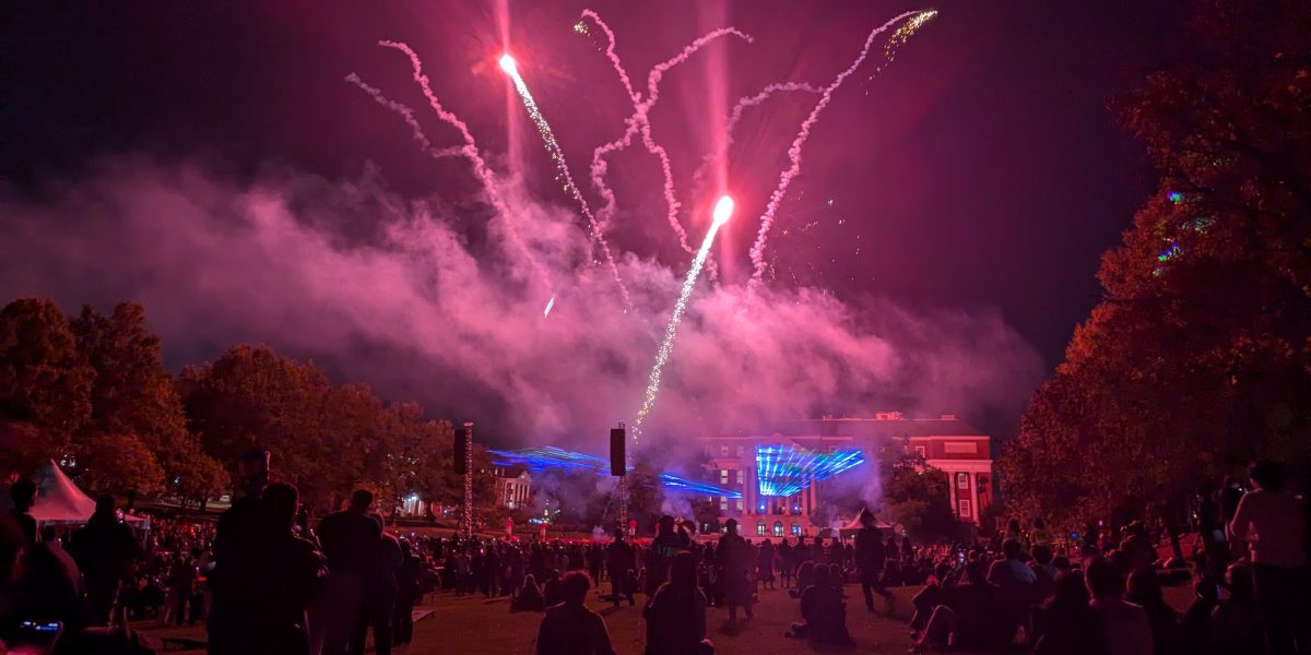 A crowd of people sits on a grassy lawn at night watching a fireworks and laser light show. Red fireworks burst in the sky while blue laser beams shine across the front of a lit-up campus building, with trees framing the scene.
