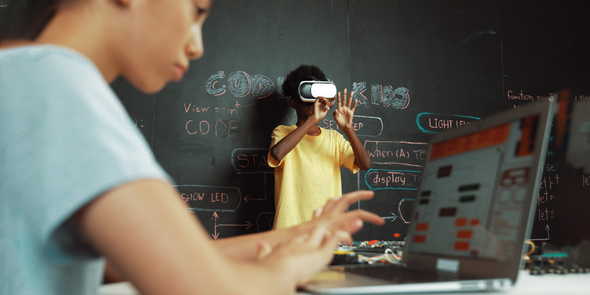A student wearing a virtual reality headset stands in front of a chalkboard covered with coding notes, while another student in the foreground types on a laptop during a classroom technology activity.