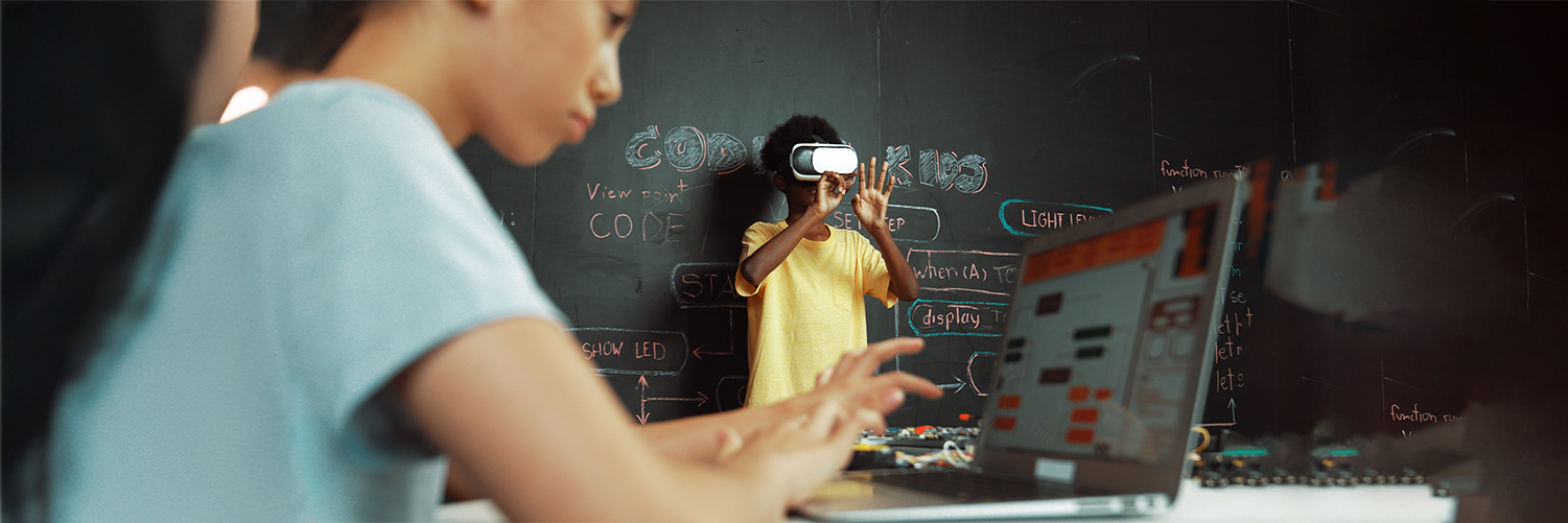 A student in the foreground works on a laptop showing code, while another young student in the background wears a virtual reality (VR) headset in front of a chalkboard covered in code and programming terms.