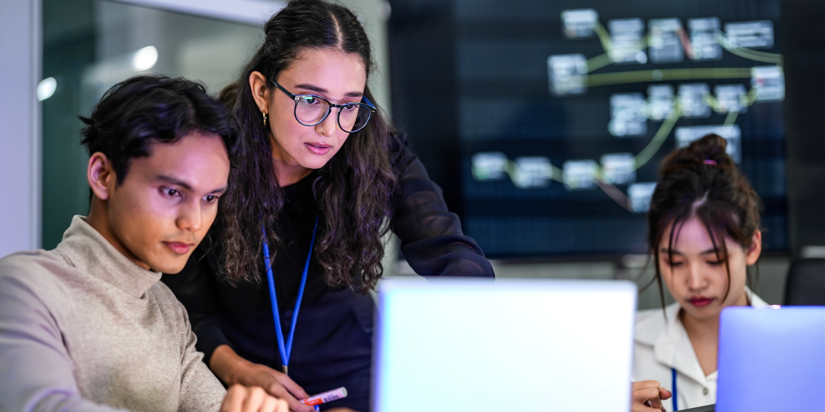 A diverse group of professionals in a corporate workshop on AI development. A female instructor supervises as the team learns to code advanced machine learning models on their laptops.