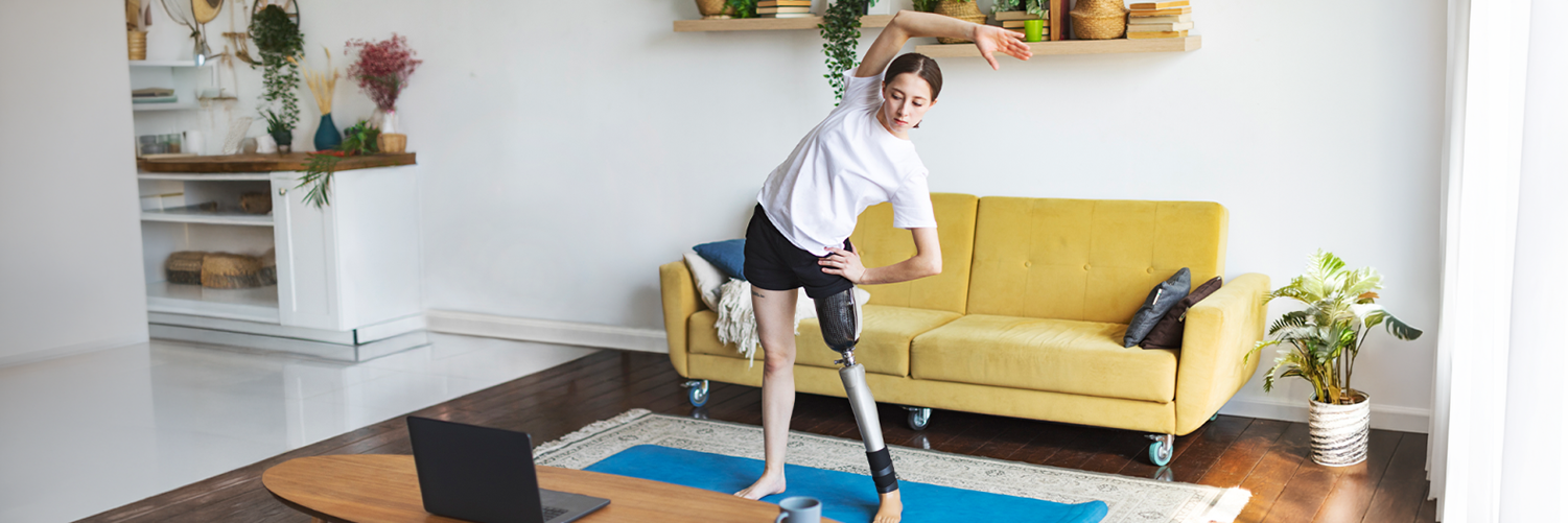 A woman with a prosthetic leg performs a side stretch on a blue yoga mat in a bright living room while following a workout on a laptop placed on a nearby coffee table.