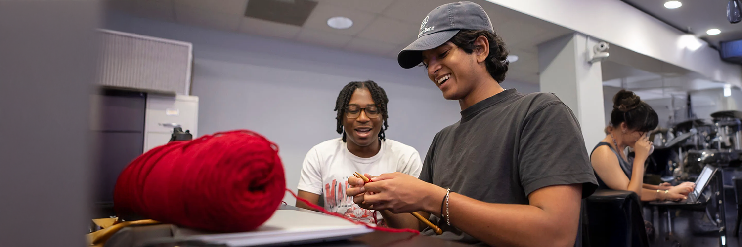 Two young men sit at a desk in a common area or classroom. One man, wearing a baseball cap and dark t-shirt, smiles as he uses wooden needles to knit with bright red yarn. His companion, wearing glasses and a white t-shirt, looks on with an encouraging smile. A large ball of red yarn sits prominently on the desk in the foreground.