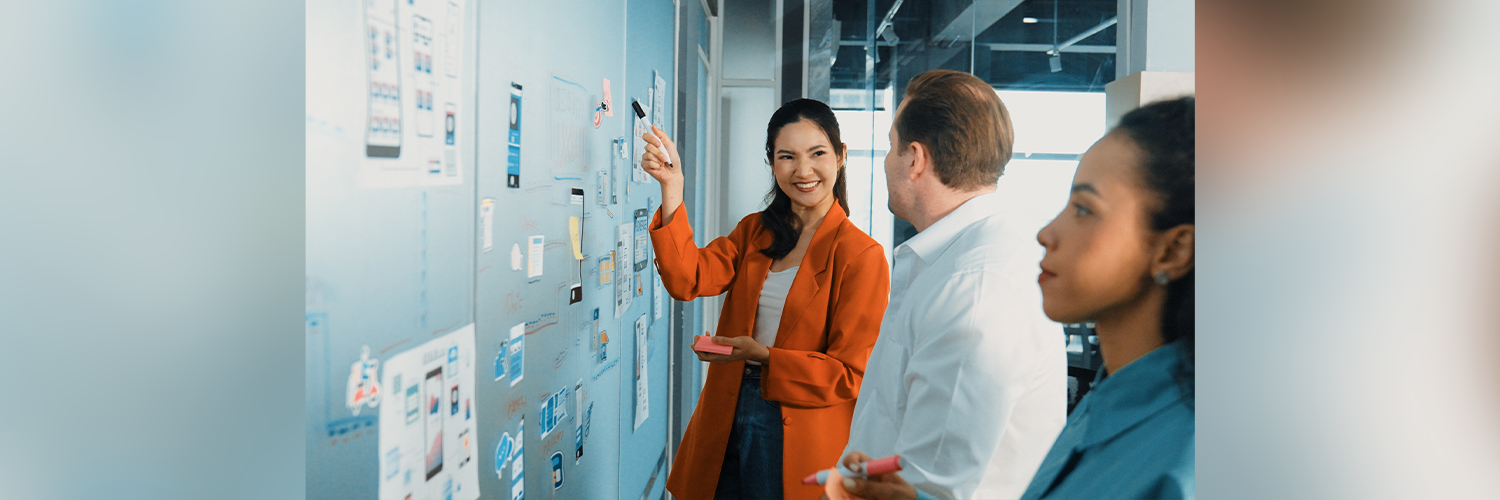 A group of diverse professionals, including a smiling Asian woman in a bright orange blazer, collaborate in an office, using sticky notes to create a user flow or wireframe on a glass wall.