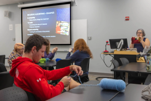 A college classroom shows a man knitting with blue yarn in the foreground, facing a screen displaying "RECURSIVE ALGORITHMS - EXAMPLES." Other students are visible, and a professor holds up knitted fabric.