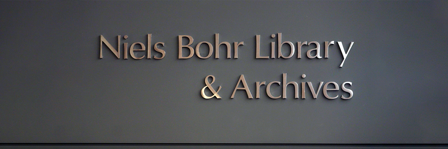 Three-dimensional, metallic copper-colored lettering mounted on a dark gray wall that reads: "Niels Bohr Library & Archives." The image is cropped in a wide, panoramic aspect ratio.