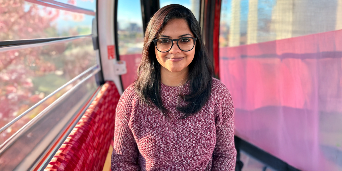 Head shot of HCIM alum Kaush Ganesh Portrait of a woman wearing glasses and a maroon sweater, standing inside a gondola with soft pink and red tones and a blurred cityscape outside.
