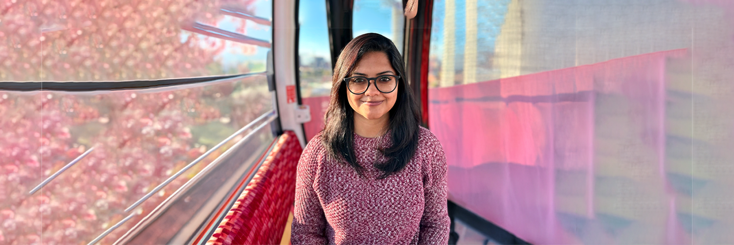 Head shot of HCIM alum Kaush Ganesh Portrait of a woman wearing glasses and a maroon sweater, standing inside a gondola with soft pink and red tones and a blurred cityscape outside.
