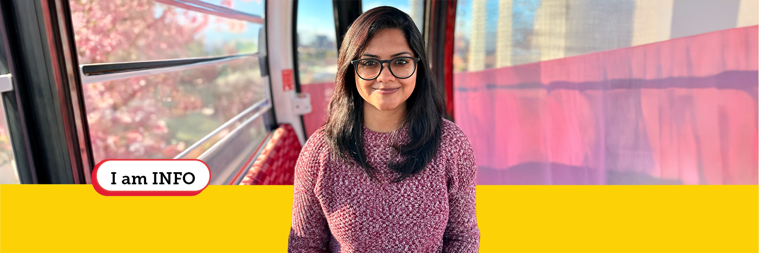 Head shot of HCIM alum Kaush Ganesh Portrait of a woman wearing glasses and a maroon sweater, standing inside a gondola with soft pink and red tones and a blurred cityscape outside.