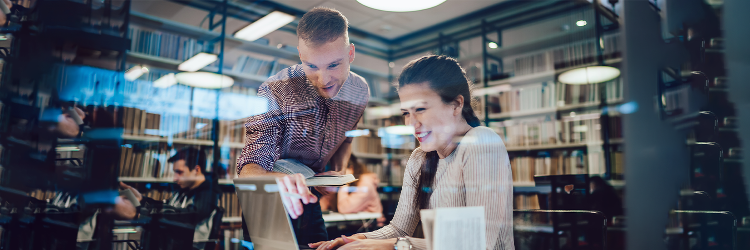 A librarian and graduate student collaborate over a laptop in a library, surrounded by bookshelves.
