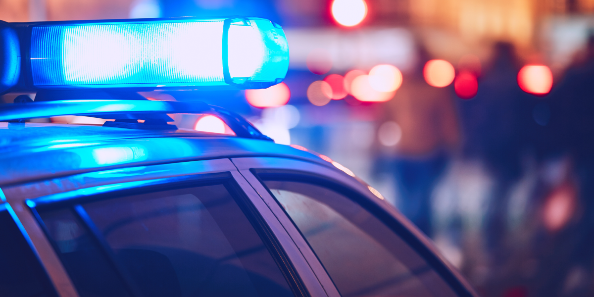 Close-up of the blue flashing emergency light bar on the roof of a police vehicle at night, with blurry red traffic lights and silhouettes of people in the background.