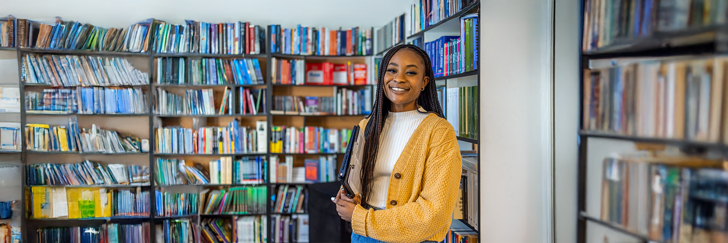 Smiling woman holding a book stands between tall library shelves filled with books, suggesting an academic or learning environment.