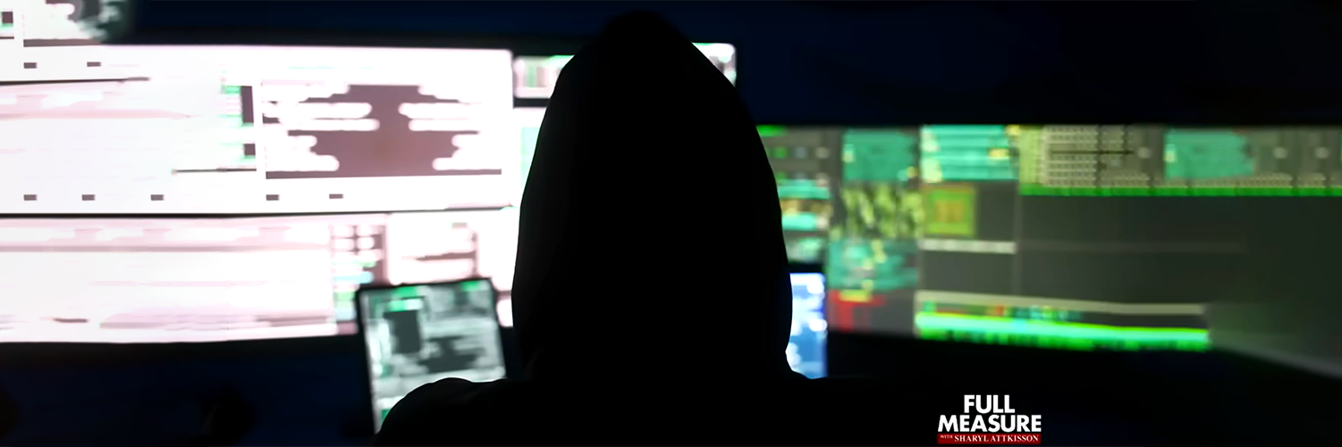 A person in a dark hoodie, seen from behind as a silhouette, working in front of multiple bright computer screens displaying code or data. The image suggests hacking or a dark cyber environment.