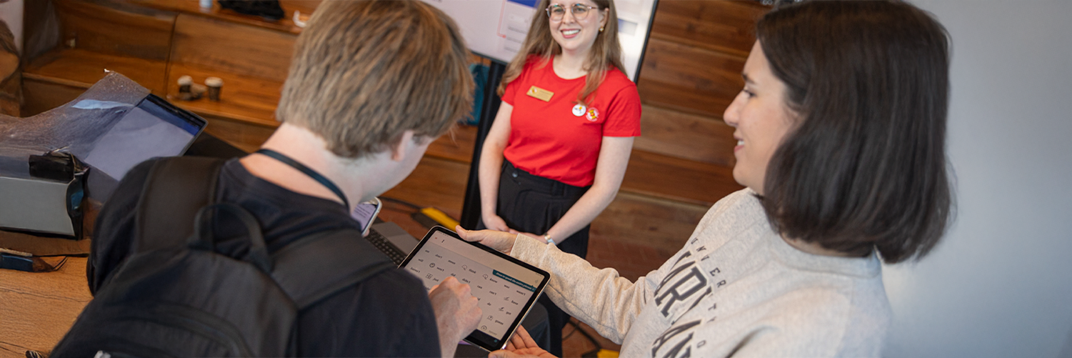 Two students, a male with a backpack and a female, look at a tablet together. Another smiling woman in a red shirt stands in the background.