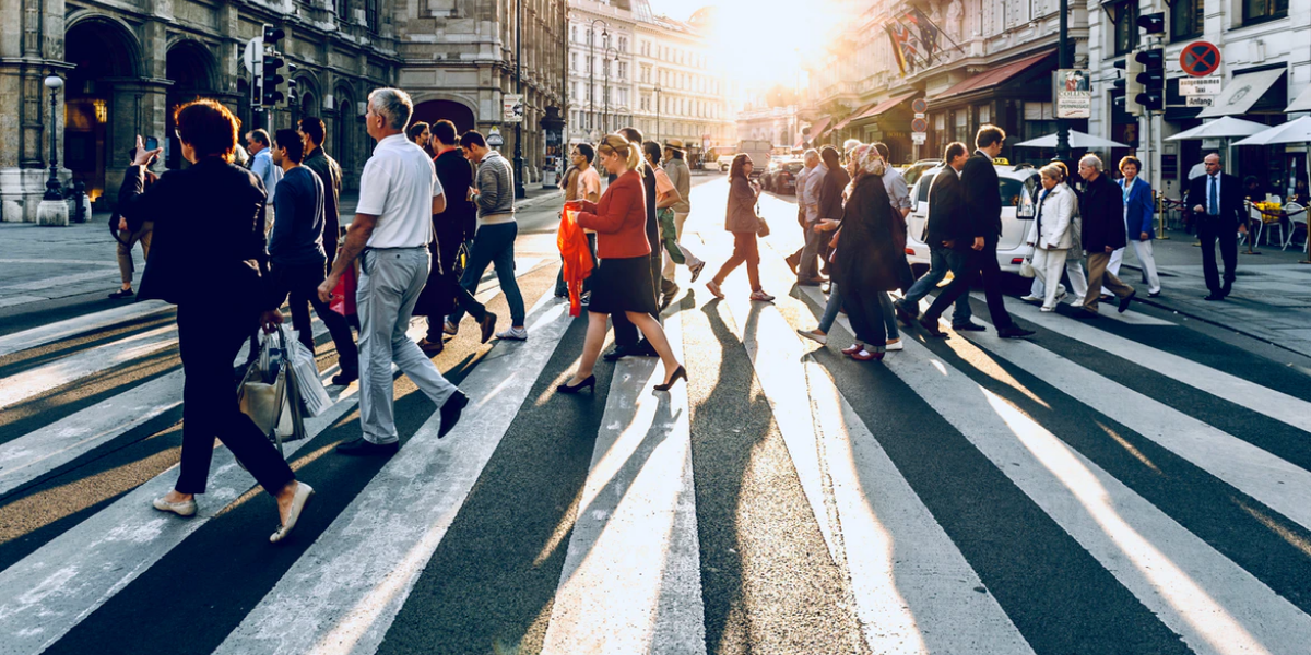 A crowd of people walking on a crosswalk with the sun setting in the background.