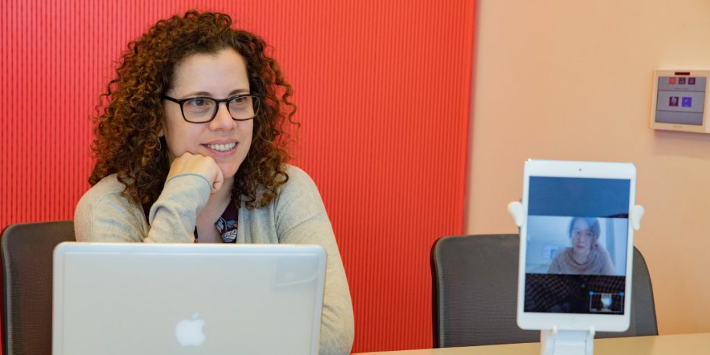 photo of Dr. Susannah Paletz sitting at a table with a laptop and iPad facing the viewer that is displaying another woman