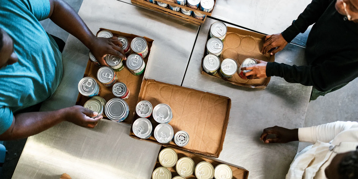 Overhead view of volunteers organizing canned goods into cardboard boxes on a metal table at a food pantry.