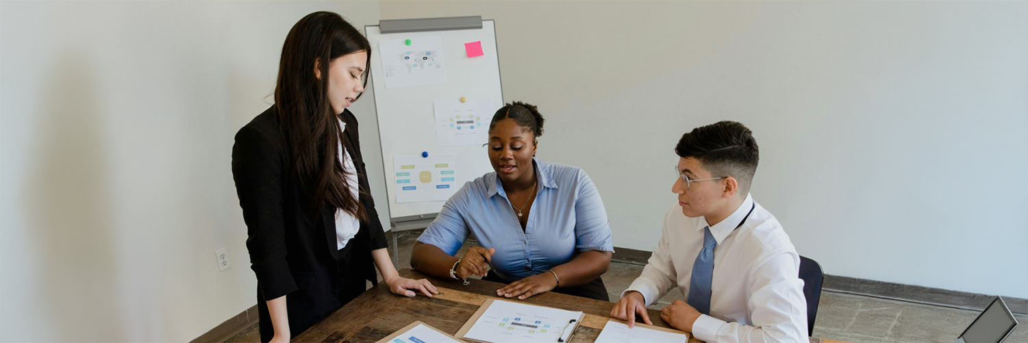 IConsultancy at INFO Three professionals collaborate at a table in a meeting room. One person stands while two others sit, reviewing printed charts and documents. A whiteboard with diagrams and sticky notes is in the background, suggesting a discussion or planning session.