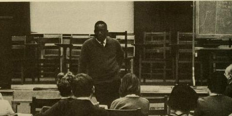 black and white photo of man standing in the front of a room with people faced towards him in chairs